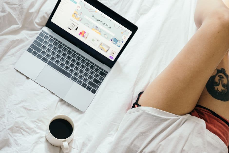 From above of unrecognizable woman with tattoo on leg browsing netbook and drinking coffee while resting on bed in cozy